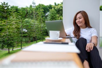 A woman covered by digital nomad insurance enjoys remote work on her laptop, sitting on a balcony with a lush green view.