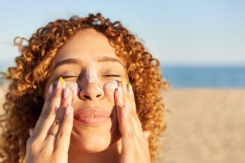a smiling happy woman applying sunscreen to her face on the beach, having read our expert skin care tips for living abroad