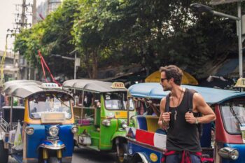 a male tourist standing next to tuk-tuks in Thailand, aware of potential travel scams like overcharging tourists in popular destinations