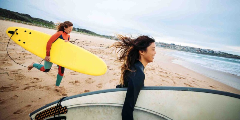 two women carrying surfboard on a beach, heading towards the sea